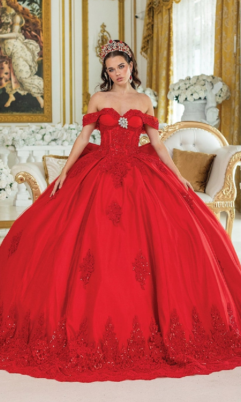 A woman wears the Quinceanera Dress 1913 By Dancing Queen, an ornate red off-the-shoulder gown, in a lavish gold-accented room with white flowers and elegant furniture, complemented by a jeweled headpiece.